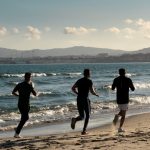 Three men jogging along a scenic beach, embracing a healthy lifestyle under a clear sky.